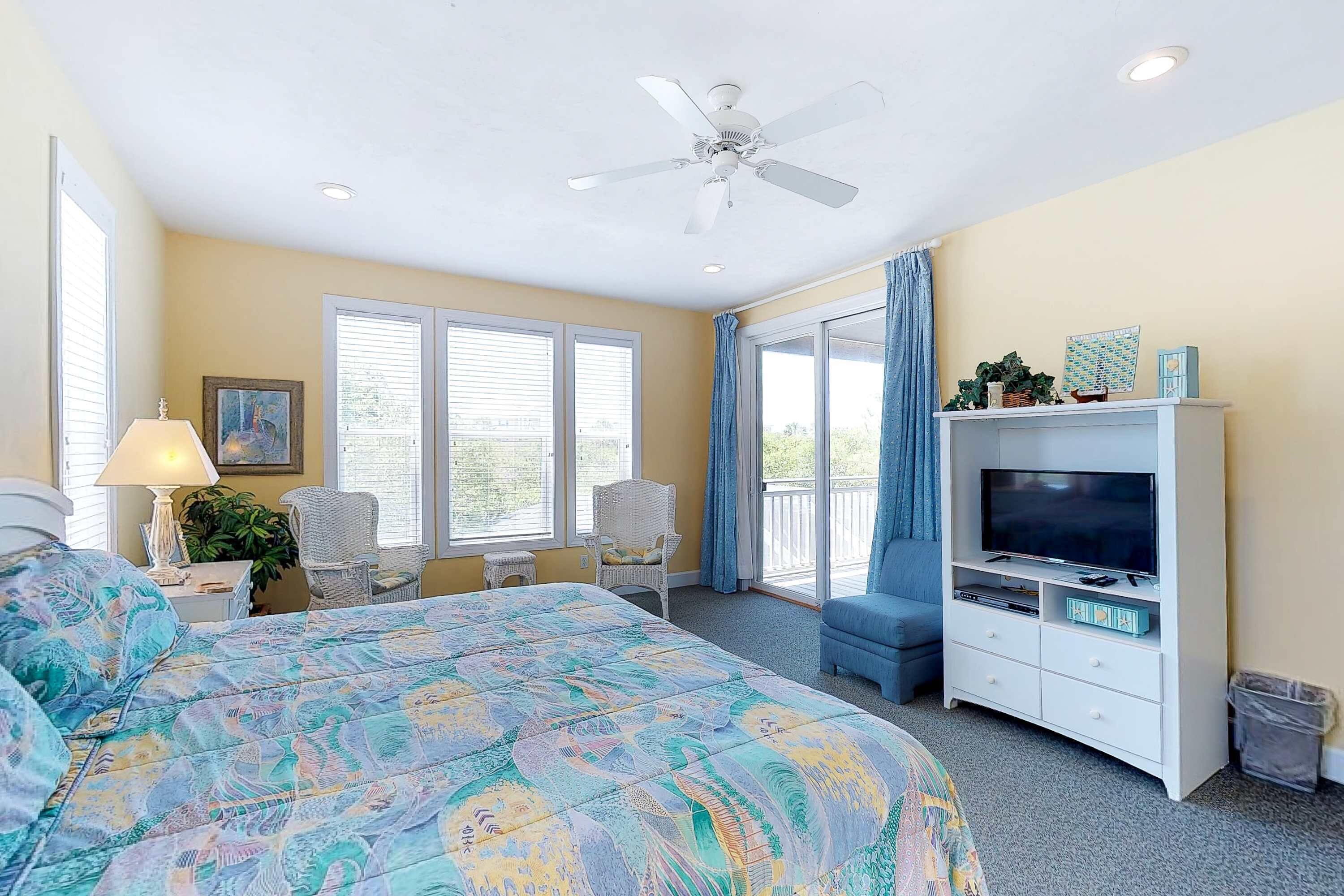 Quarto com cama, móvel para televisão, ventilador de teto e grandes janelas.