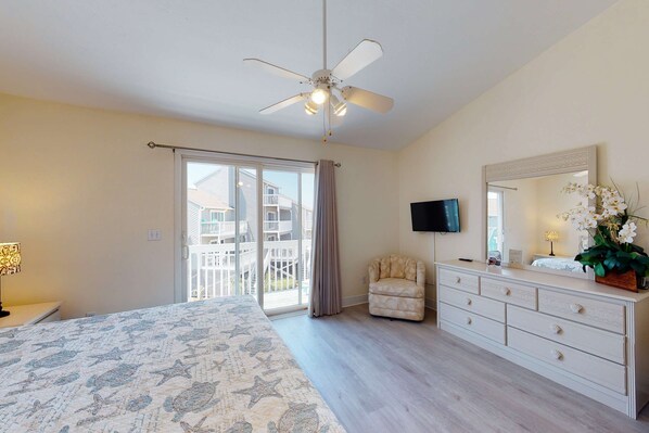 A bedroom with a bed, a dresser, a chair, a ceiling fan, a TV, and a balcony with a view of other buildings. - Dog-friendly townhome in private community with pool - near beach (Cape San Blas)