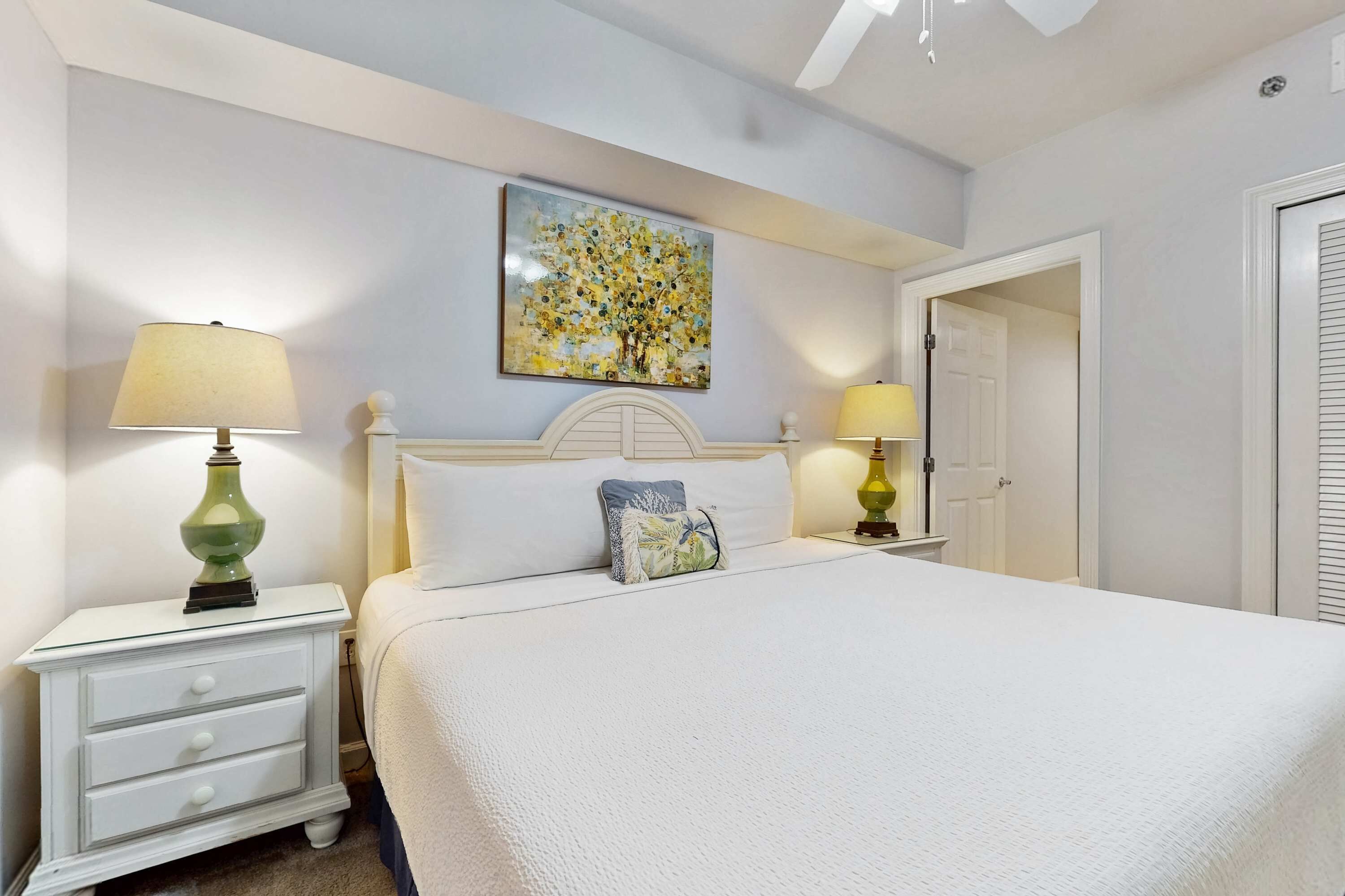A neatly arranged bedroom with a large bed, two bedside tables with lamps, a decorative pillow, and a wall painting.