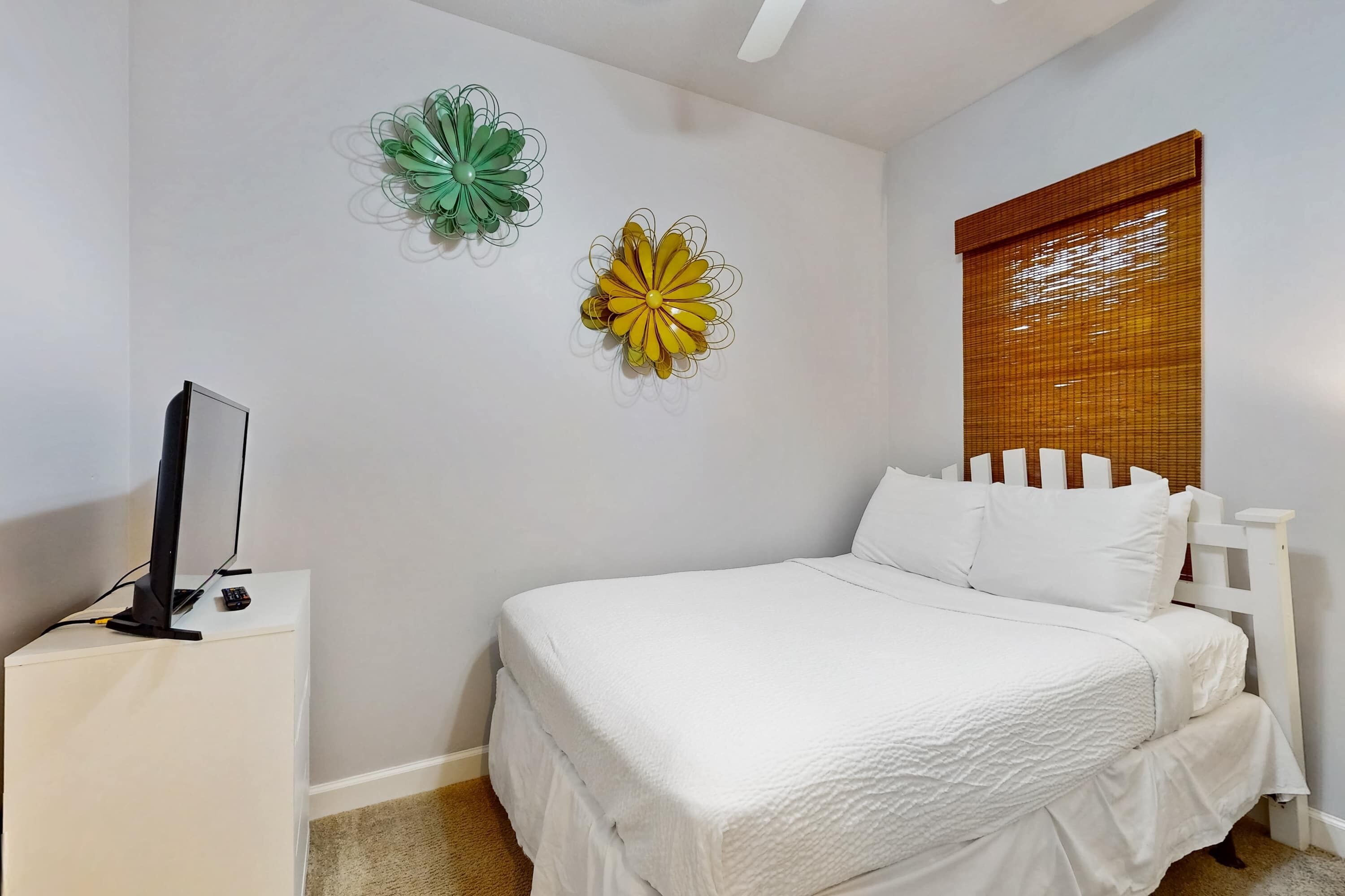 Une chambre avec un lit blanc, une table de chevet blanche sur laquelle se trouve un téléviseur, et des œuvres d’art décoratives accrochées au mur.