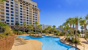 A swimming pool with a curved design, surrounded by palm trees and lounge chairs, with a multi-story building in the background. - Dreamy coastal condo w/ ocean view on-site pool, hot tub & easy beach access (Miramar Beach)