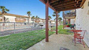 A covered patio area with a concrete floor, red metal chairs, and a small table. There are palm trees and a fenced area in the background. - Pool-side condo with pool, private washer&dryer, walk to beach & free WiFi (Navarre Beach)
