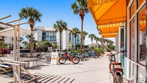 A sunny street with bicycles parked along the sidewalk, palm trees, and a building with a yellow awning. - Bright condo near shopping & restaurants with tennis & shared pools (Santa Rosa Beach)