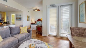 A spacious living room with a gray sofa, a marble coffee table, and a wooden bench. A ceiling fan and a large window with blinds are visible. - Bright condo near shopping & restaurants with tennis & shared pools (Santa Rosa Beach)