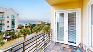 A balcony with a view of the ocean, a white door with blinds, and a street with palm trees. - Bright condo near shopping & restaurants with tennis & shared pools (Santa Rosa Beach)
