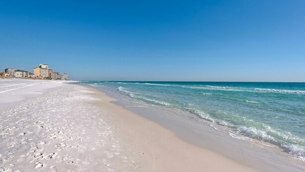 Ein Strand mit blauem Himmel, sandigem Ufer und Gebäuden im Hintergrund.