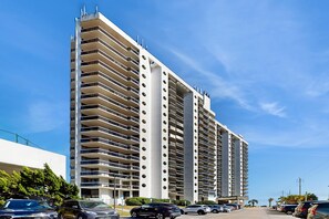 A high-rise building with multiple balconies, a parking lot with several cars, and a clear blue sky. - Cozy escape with Gulf views from balcony, pool, hot tub & WD - access to beach (Miramar Beach)