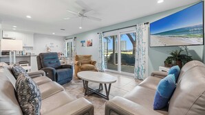 A spacious living area with a ceiling fan, a large TV screen, and a view of the beach through the sliding glass doors. - Sunny condo with lake views, outdoor pool, fast WiFi, tennis & central AC (Miramar Beach)