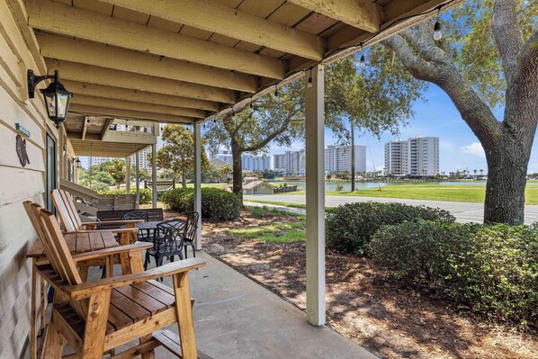A covered outdoor seating area with wooden chairs and tables, a pergola overhead, and a view of a residential area with buildings and a tree. - Sunny condo with lake views, outdoor pool, fast WiFi, tennis & central AC (Miramar Beach)