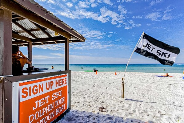 Ein Strand mit einem Schild für Jetskis, einer Anmeldetafel für eine Delfintour und einem strahlend blauen Himmel.