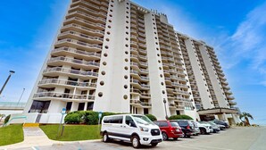 A high-rise building with multiple balconies and a parking lot with several cars. - Gorgeous ground-floor condo, private patio,  shared pool and beach access (Miramar Beach)