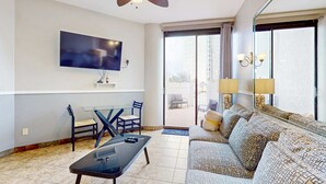 A modern living room with a flat-screen TV, a ceiling fan, a sofa, a coffee table, and a dining area with chairs. - Gorgeous ground-floor condo, private patio,  shared pool and beach access (Miramar Beach)