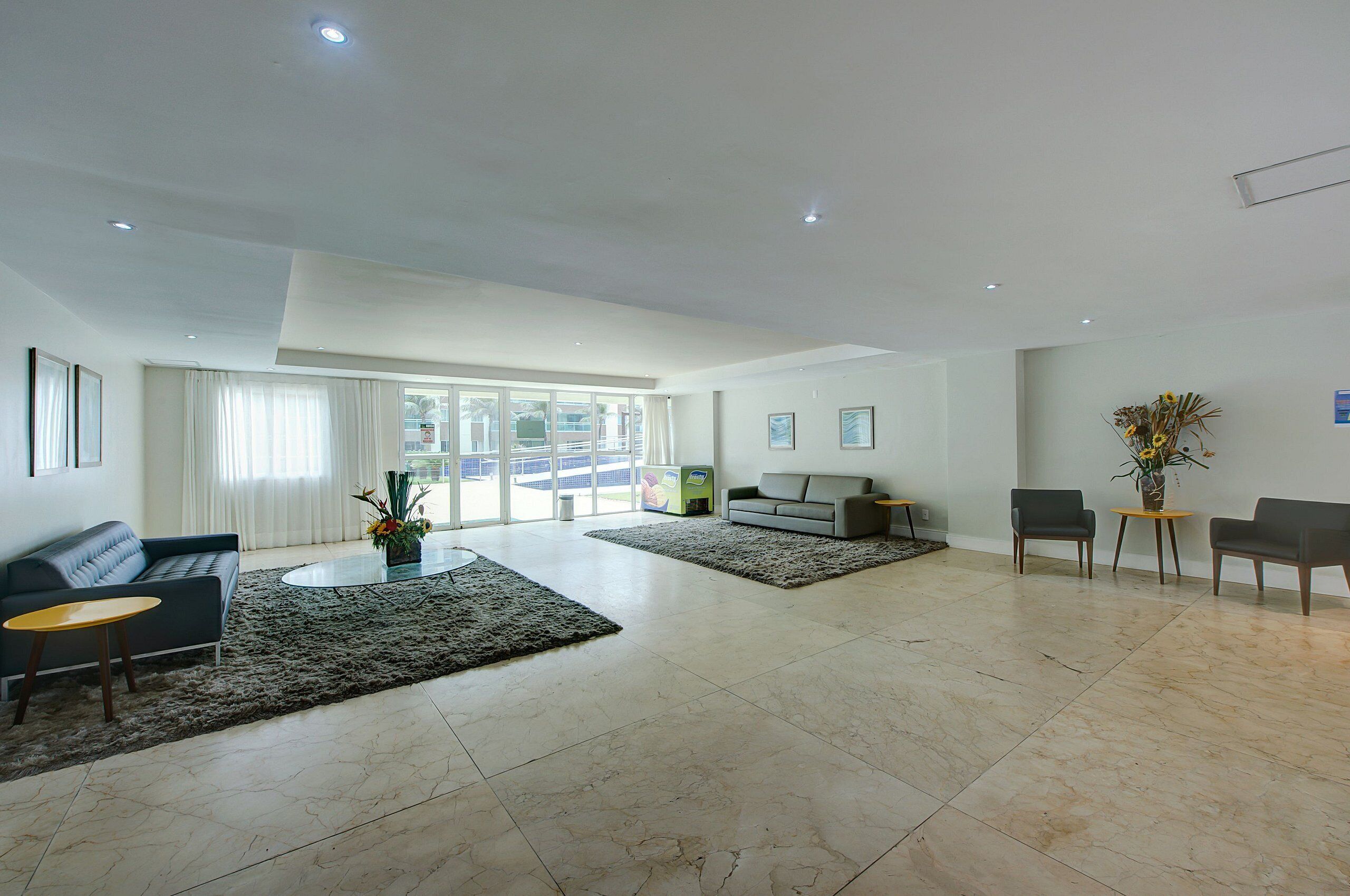 A spacious living room with a grey sofa, a coffee table, and a TV. There are two chairs and a small table with a vase of flowers. The room has large windows and a view of the outside.