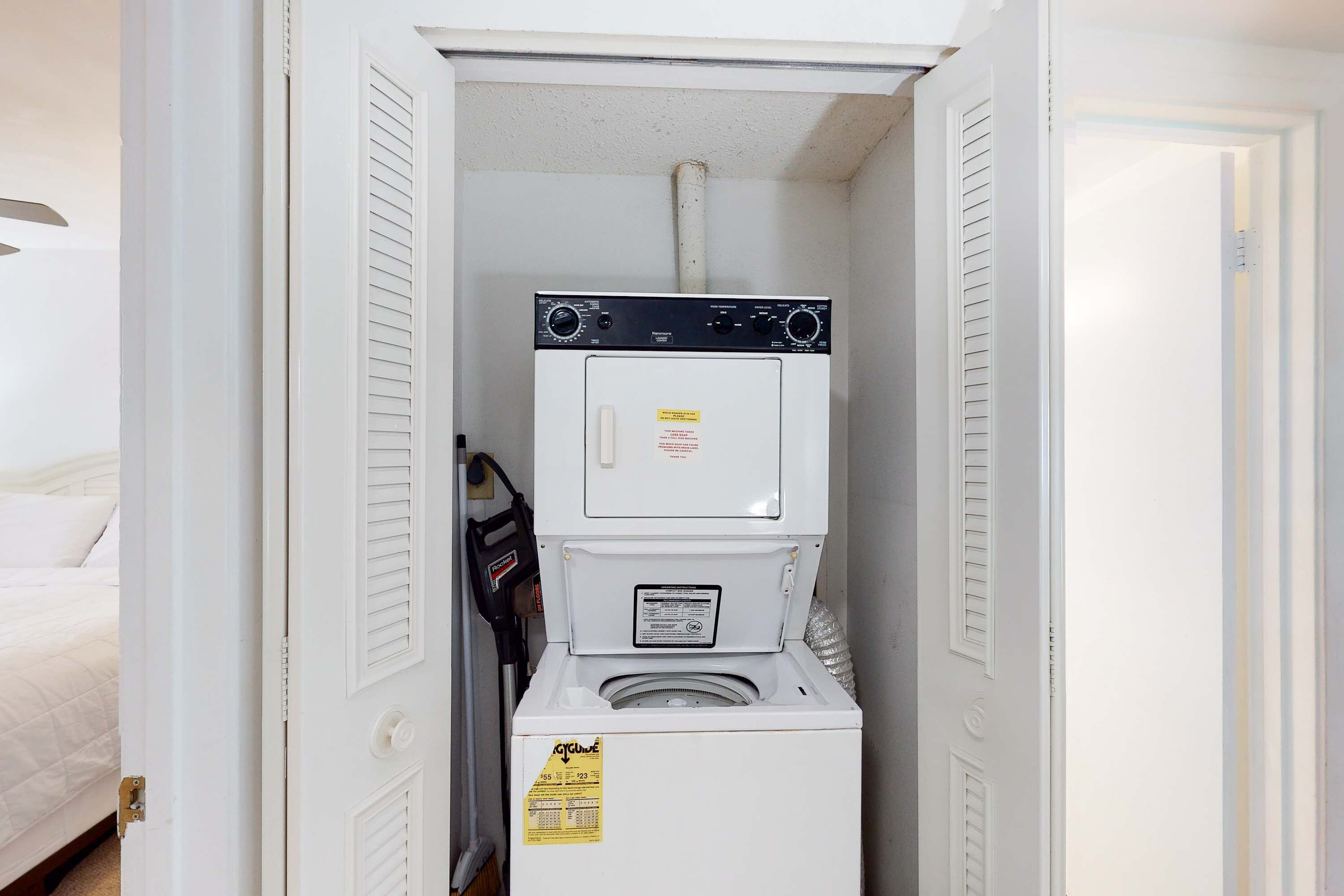 A compact laundry room with a front-loading washer and dryer, a small shelf, and a door leading to a bedroom.