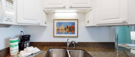 A kitchen with white cabinets, a granite countertop, a double sink, a coffee maker, paper towels, and a framed painting above the sink.