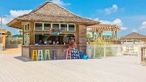 A wooden beachside bar with a thatched roof, a signboard, and colorful stools. - Gulf-view condo at beachfront resort with pools, hot tub & 2 gyms (Destin)