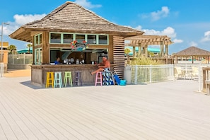 A wooden beachside bar with a thatched roof, a signboard, and colorful stools. - Gulf-view condo at beachfront resort with pools, hot tub & 2 gyms (Destin)
