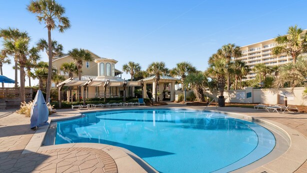 A swimming pool with a tiled deck, surrounded by palm trees and a building with a light-colored facade. - Contemporary condo near the beach,  Shared pool & gym (Destin)