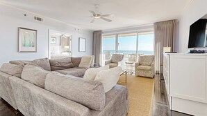 Een ruime woonkamer met een grote beige hoekbank, een flatscreen televisie op een wit kastje, en een balkon met buitenmeubels.