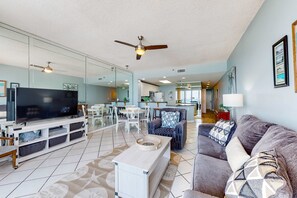A spacious living room with a flat-screen TV, a ceiling fan, and a dining area in the background. - Cozy condo with Gulf-front balcony & great views - near shops & dining (Destin)