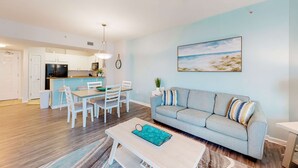 A modern living room with a light blue sofa, a dining table with chairs, and a kitchen area in the background. - Beachy resort condo with balcony, outdoor pools, hot tub, gym, tennis, AC, & W&D (Destin)