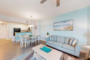 A modern living room with a light blue sofa, a dining table with chairs, and a kitchen area in the background. - Beachy resort condo with balcony, outdoor pools, hot tub, gym, tennis, AC, & W&D (Destin)