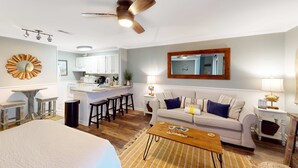 Ein modernes Wohnzimmer mit einer Couch, einem Couchtisch und einem Deckenventilator. Im Hintergrund ist eine Küche zu sehen.