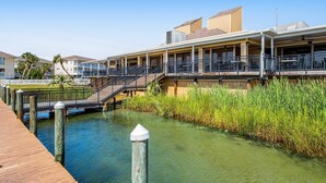 Ein Hotel am Wasserufer mit einer Holzterrasse, einer Brücke und einem Rasen mit hohen Schilfhalmen.