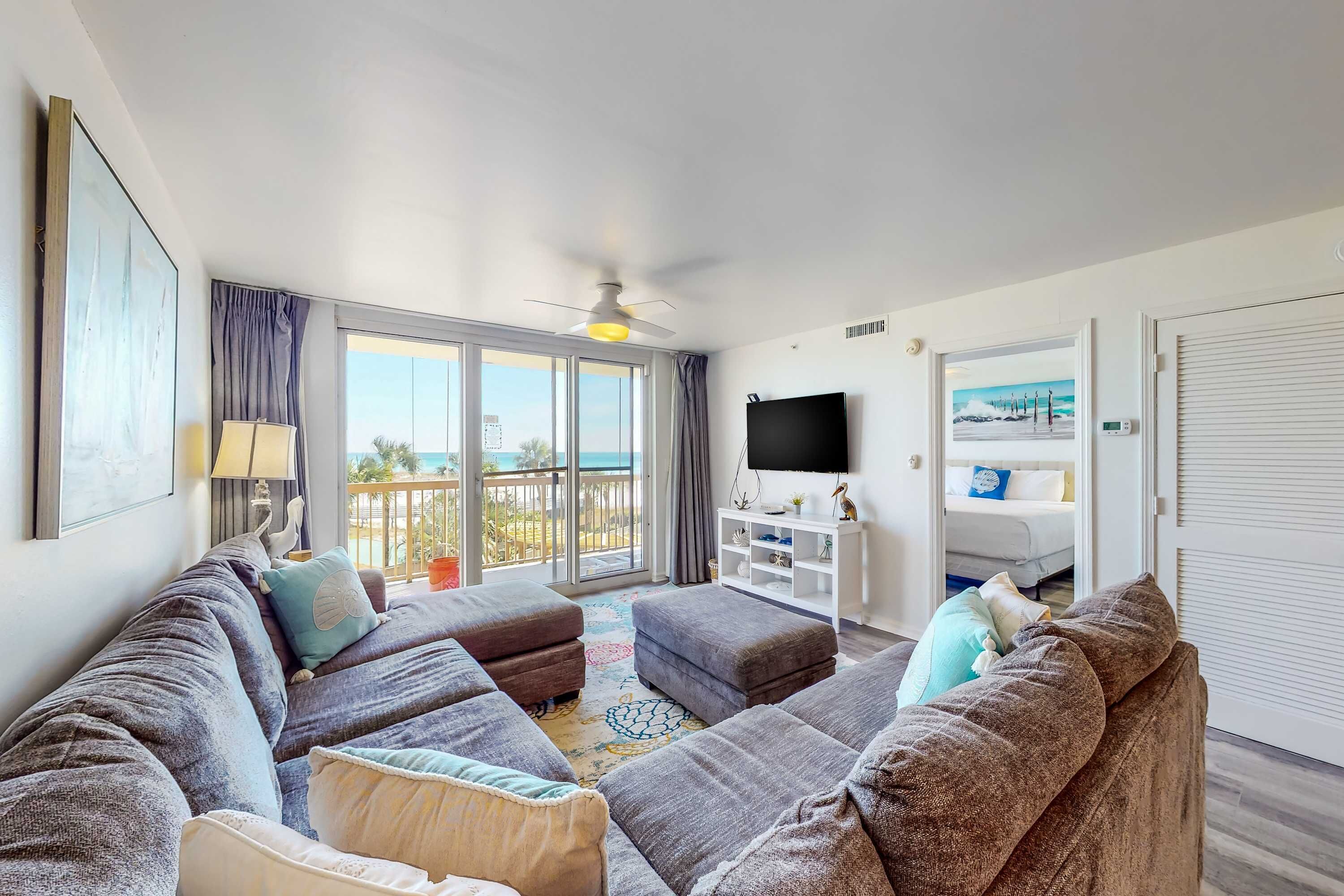 A spacious living room with a large gray sectional sofa, a flat-screen TV, and a balcony with a view of the ocean.