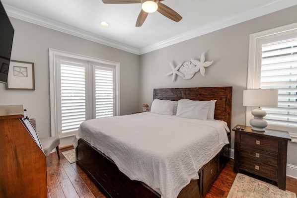A bedroom with a large bed, wooden headboard, white bedding, a nightstand with a lamp, and a ceiling fan. - Villa Lago - Emerald Isle (Miramar Beach)