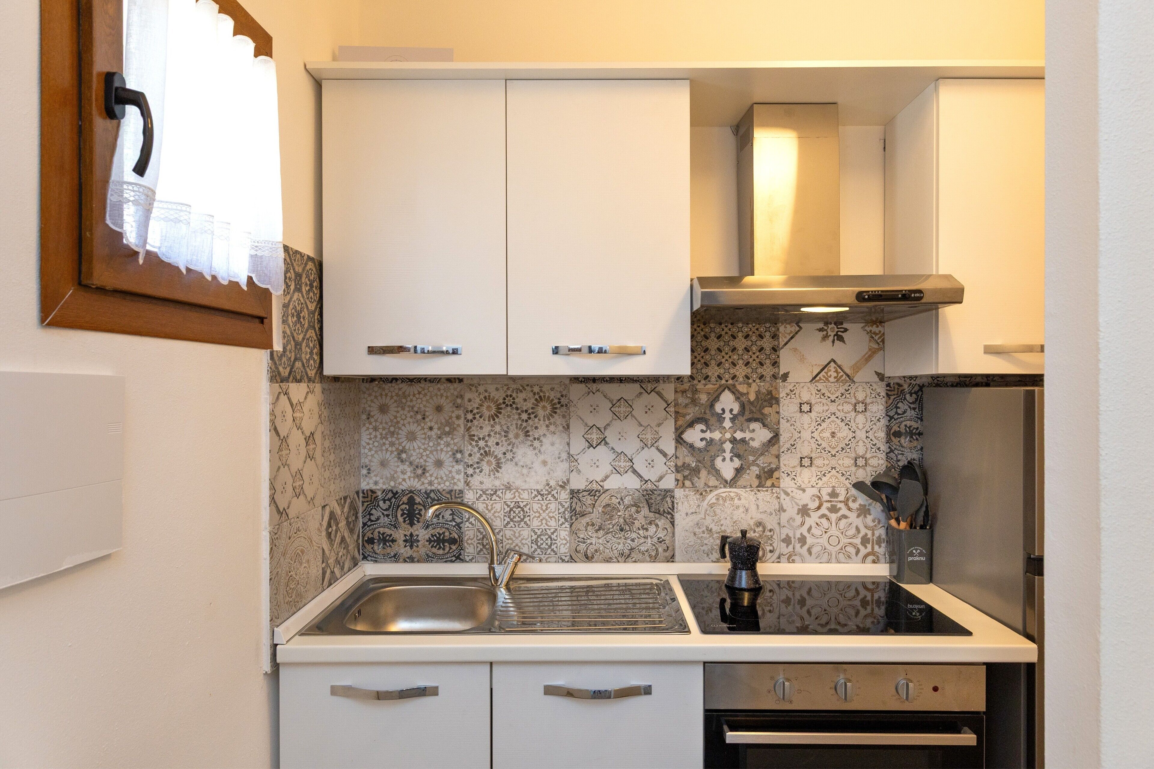 Una cocina compacta con armarios blancos, un lavamanos de acero inoxidable y azulejos decorativos en la pared.
