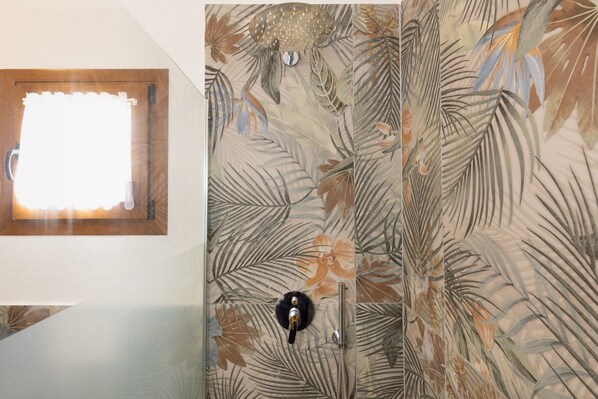 A bathroom with a shower, a window with a curtain, and a wall with a tropical leaf pattern.