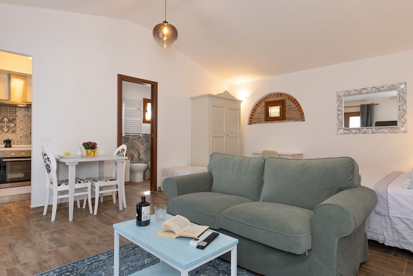 A living room with a green sofa, a white table with a remote and book, a dining area with chairs, and a kitchen visible in the background. - Apartment 'Il Mulino Del Botteghino - A' with Shared Garden, Wi-Fi & Air Conditioning (Botteghino)