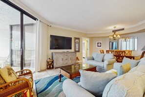 A living room with a sofa, armchair, and a glass coffee table. There is a television on a cabinet, and a chandelier hangs from the ceiling. - Gorgeous beachside condo with pool and easy beach access (Miramar Beach)