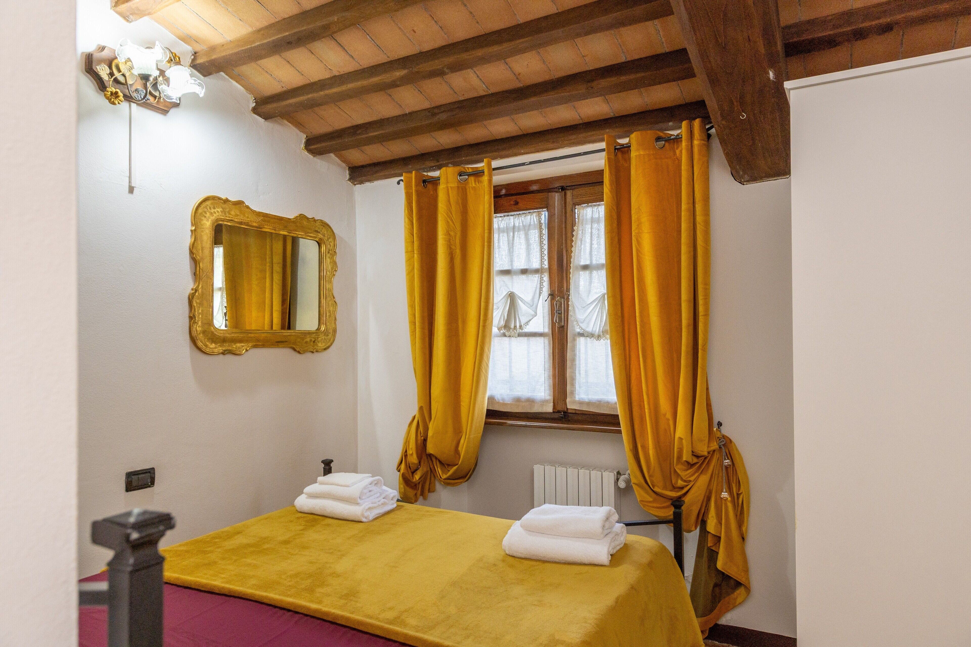Une chambre avec un plafond en bois, un lit recouvert d’une couverture jaune, un miroir avec un cadre doré et des rideaux jaunes à la fenêtre.