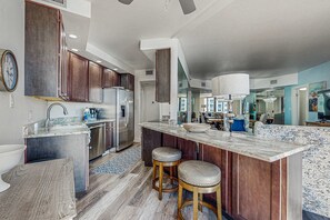 A modern kitchen with dark wooden cabinets, stainless steel appliances, and a marble island with stools. - Beach + Ocean View Condo with WiFi, Private Balcony, AC, & Pool/Hot Tub/Tennis (Miramar Beach)