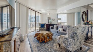 Uma sala de estar ampla com uma janela grande, uma mesa de centro de madeira, uma poltrona estampada e um sofá.