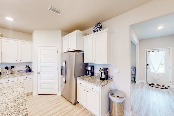 A modern kitchen with white cabinetry, a stainless steel refrigerator, granite countertops, and a coffee maker. - Brand-new beach house with pool, beach access, AC, & W/D, Snowbirds welcome! (Port St. Joe)