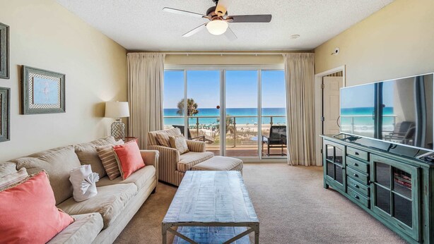A hotel room with a large window offering a view of the beach, a ceiling fan, a television, and a seating area with a sofa and a coffee table. - Stunning, waterfront home with Gulf views, pool & fitness room access (Destin)