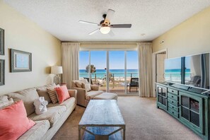 Ein Hotelzimmer mit großem Fenster und Blick auf den Strand, Deckenventilator, Fernseher und einer Sitzecke mit Sofa und Couchtisch.