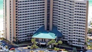 Um hotel de grande altura com vários andares, uma entrada central com telhado verde e um estacionamento com diversos carros.