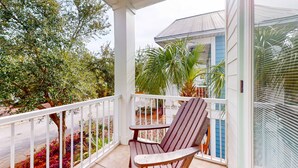 A covered balcony with a wooden chair and a small table, overlooking a garden with various plants and a building in the background. - Snowbird-friendly home in gated community with hot tub, pool, balcony, & grill (Destin)