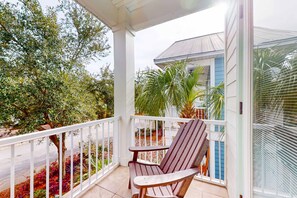 A covered balcony with a wooden chair and a small table, overlooking a garden with various plants and a building in the background. - Snowbird-friendly home in gated community with hot tub, pool, balcony, & grill (Destin)