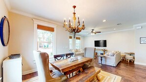 A spacious living room with a wooden dining table, comfortable seating, a chandelier, and a flat-screen TV mounted on the wall. - Snowbird-friendly home in gated community with hot tub, pool, balcony, & grill (Destin)
