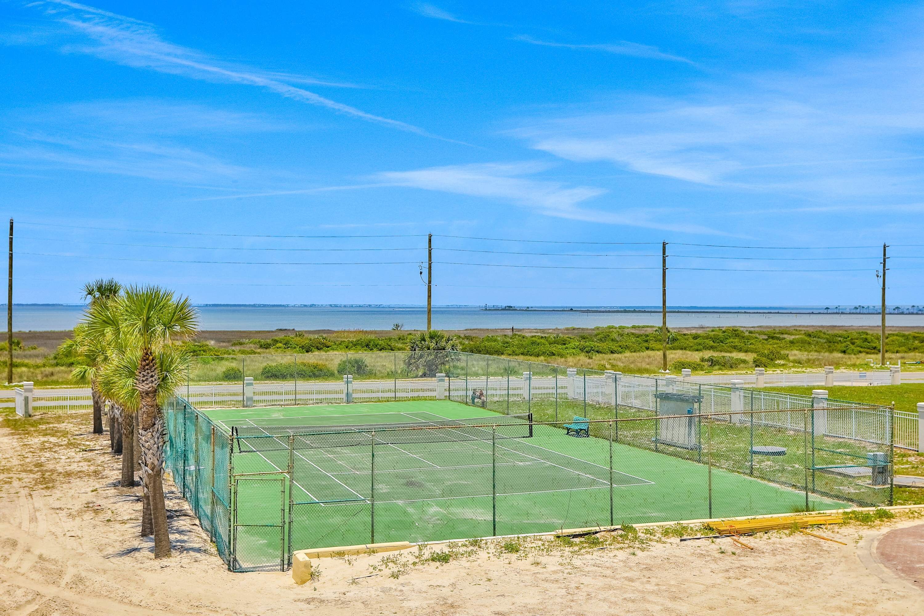 A tennis court with a net, surrounded by a fence, and palm trees in the foreground.