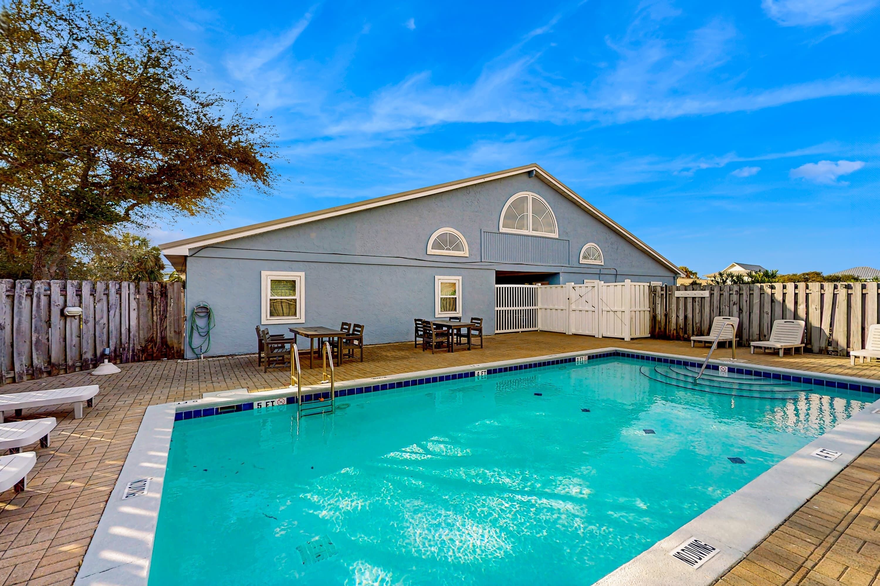 A swimming pool with a wooden deck, a covered patio area, and a house with a grey exterior and white trim.