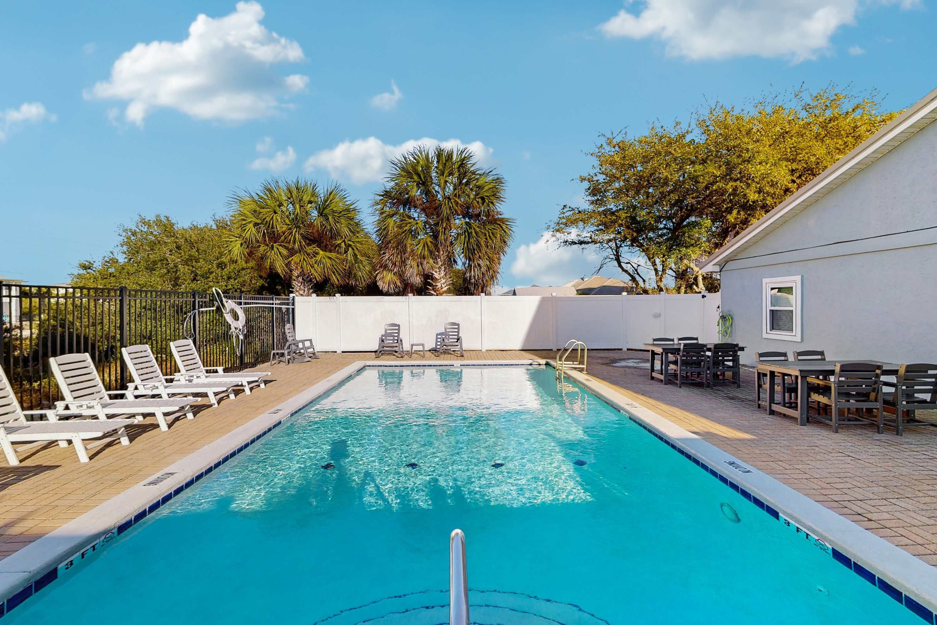 A swimming pool with lounge chairs and tables, surrounded by a white building and trees.