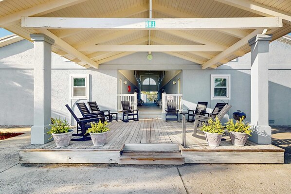 A covered outdoor seating area with wooden benches and potted plants, leading to a building entrance.