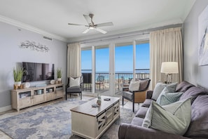 A living room with a TV, sofa, coffee table, and a balcony with a view. - Beach-view penthouse condo with stunning water views, on-site pool & hot tub (Destin)