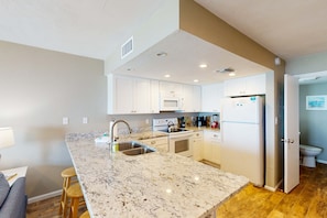 A modern kitchen with white cabinets, a granite countertop, stainless steel appliances, and a wooden stool. - Beachfront East End townhouse with beach views, deck, on-site pool & grills (St. George Island)